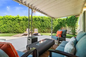 A covered patio area with outdoor furniture, a fire pit, and a view of a well-maintained hedge. - The Blue Door Bungalow - Your Private Palm Springs Home (Palm Springs)