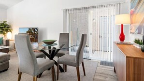 Un salon moderne avec une table à manger en verre, des chaises à revêtement gris, un lampadaire rouge et une grande fenêtre avec des stores.