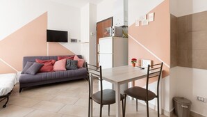 A compact living space with a sofa, a dining table with chairs, a TV mounted on the wall, a microwave, and a small trash bin. - GuestHost - Affori FN Modern Apartment x4! (Milano)