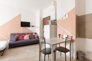 Un espace de vie compact comprenant un canapé, une table à manger avec des chaises, un téléviseur fixé au mur, un four à micro-ondes et une petite poubelle.