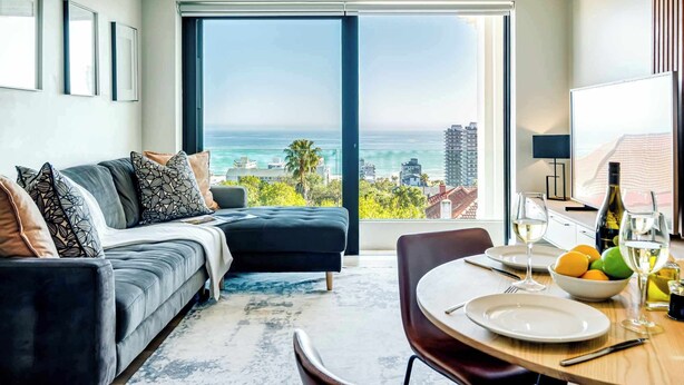 Uma sala de estar moderna com uma janela grande que oferece vista para a cidade e para o oceano, uma área de jantar com mesa posta para dois, e uma área de estar aconchegante com sofá e almofadas.