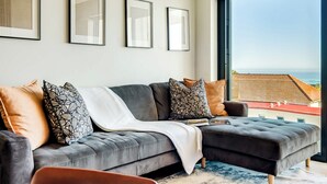 Uma sala de estar moderna com um sofá modular cinza, almofadas estampadas e uma grande janela com vista para a costa.