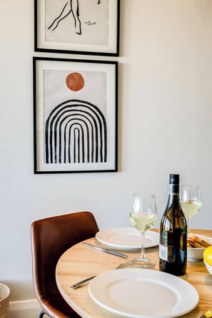 Une table à manger avec des assiettes, des verres, une bouteille de vin et une chaise. Au-dessus de la table, on peut voir deux œuvres d’art encadrées.