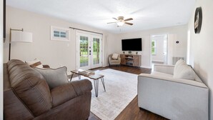 A spacious living room with a brown leather sofa, a grey armchair, a coffee table, and a flat-screen TV. There is a ceiling fan, a large window, and a door leading to an outdoor area. - Mammoth Pond - Mammoth Cave Family Retreat - Playground Sleeps 6 (Cave City)
