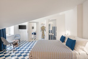 Una camera d'albergo con pavimento a scacchi, un letto con lenzuola a righe, una TV montata a parete e un bagno con uno specchio e piastrelle a righe blu.