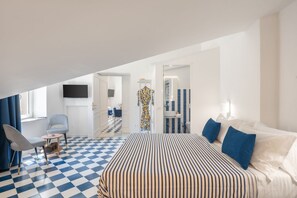 Ein Hotelzimmer mit Schachbrettmuster-Boden, einem Bett mit gestreifter Bettwäsche, einem Fernseher, einem Sessel und einem Badezimmer mit Spiegel und Waschbecken.