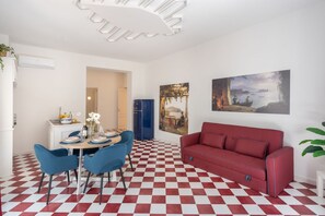 A room with a red and white checkered floor, a red sofa, a dining table with blue chairs, and two paintings on the wall.