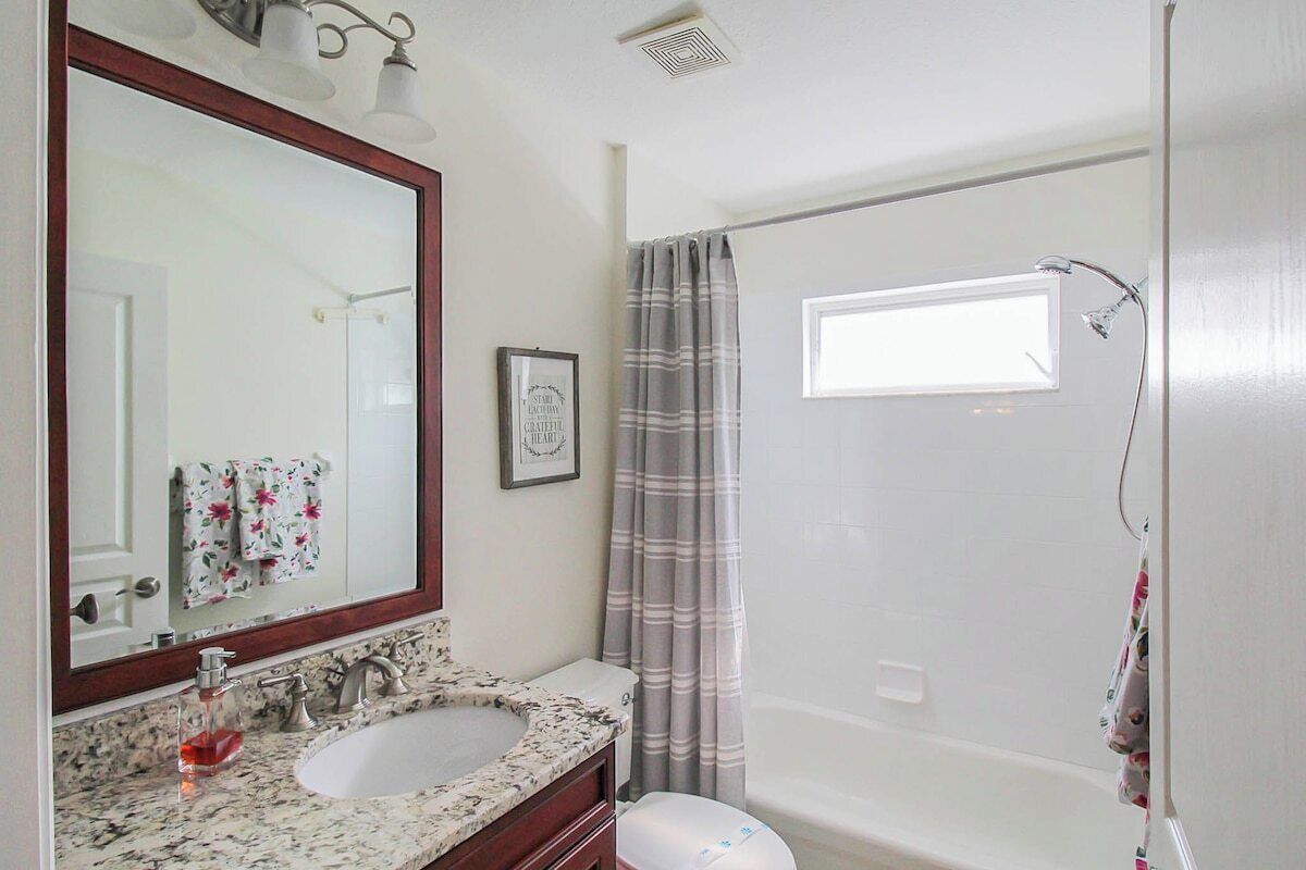 Une salle de bain avec un lavabo en marbre, un grand miroir, un tableau encadré au mur, une douche avec un rideau et une fenêtre au-dessus de la douche.