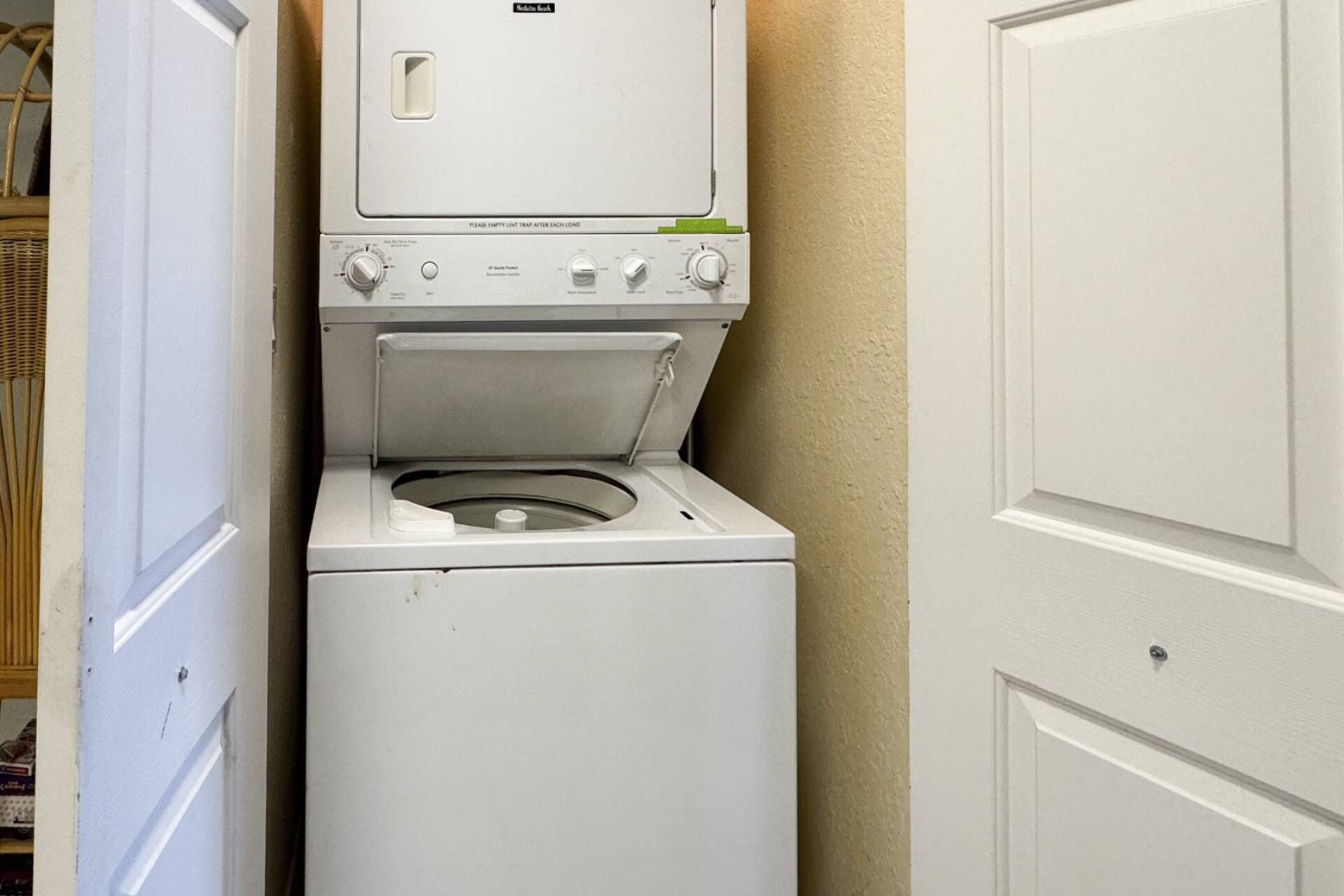 A compact, white laundry machine with a front-loading door, situated in a narrow hallway with a door on the right.