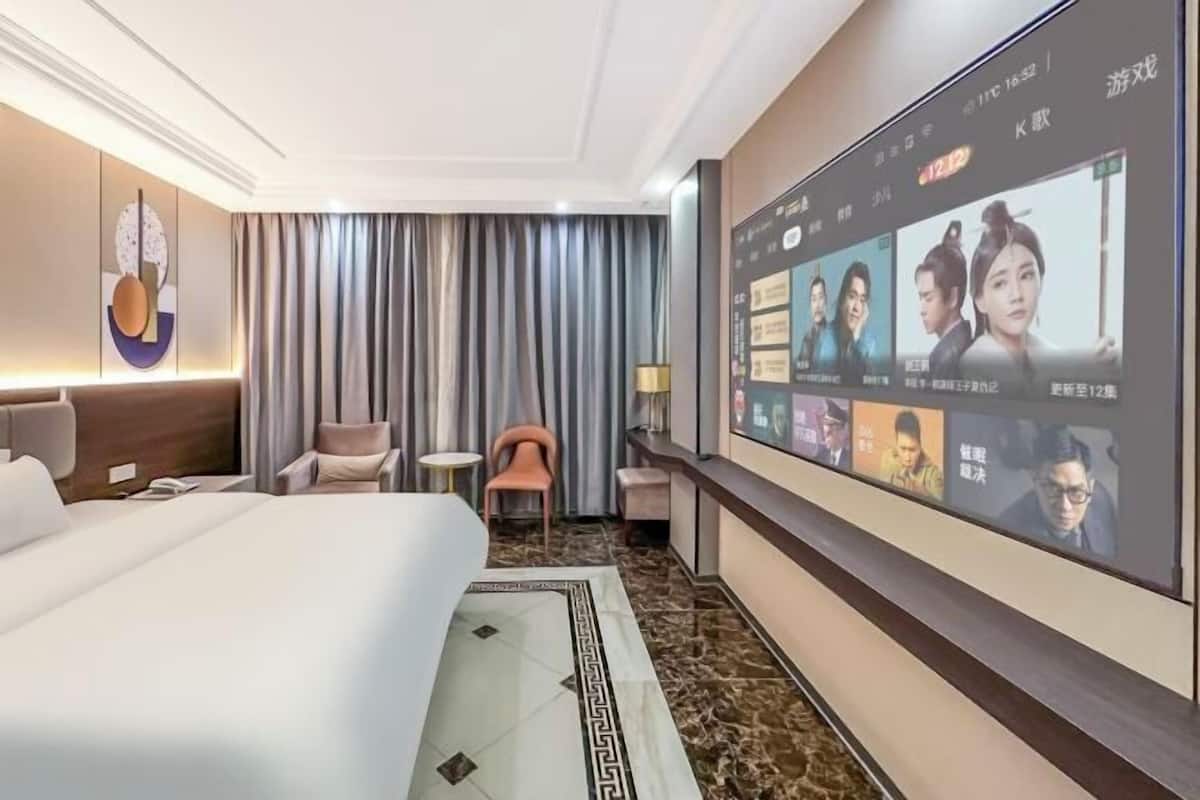 Ein Hotelzimmer mit einem großen Bett, einem an der Wand befestigten Fernseher, der ein Programm zeigt, einem kleinen Tisch mit einer Lampe und einem Sessel.