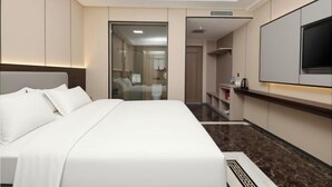 Quarto de hotel com uma cama grande, televisão de tela plana, minibar e banheiro visível através de uma porta de vidro.