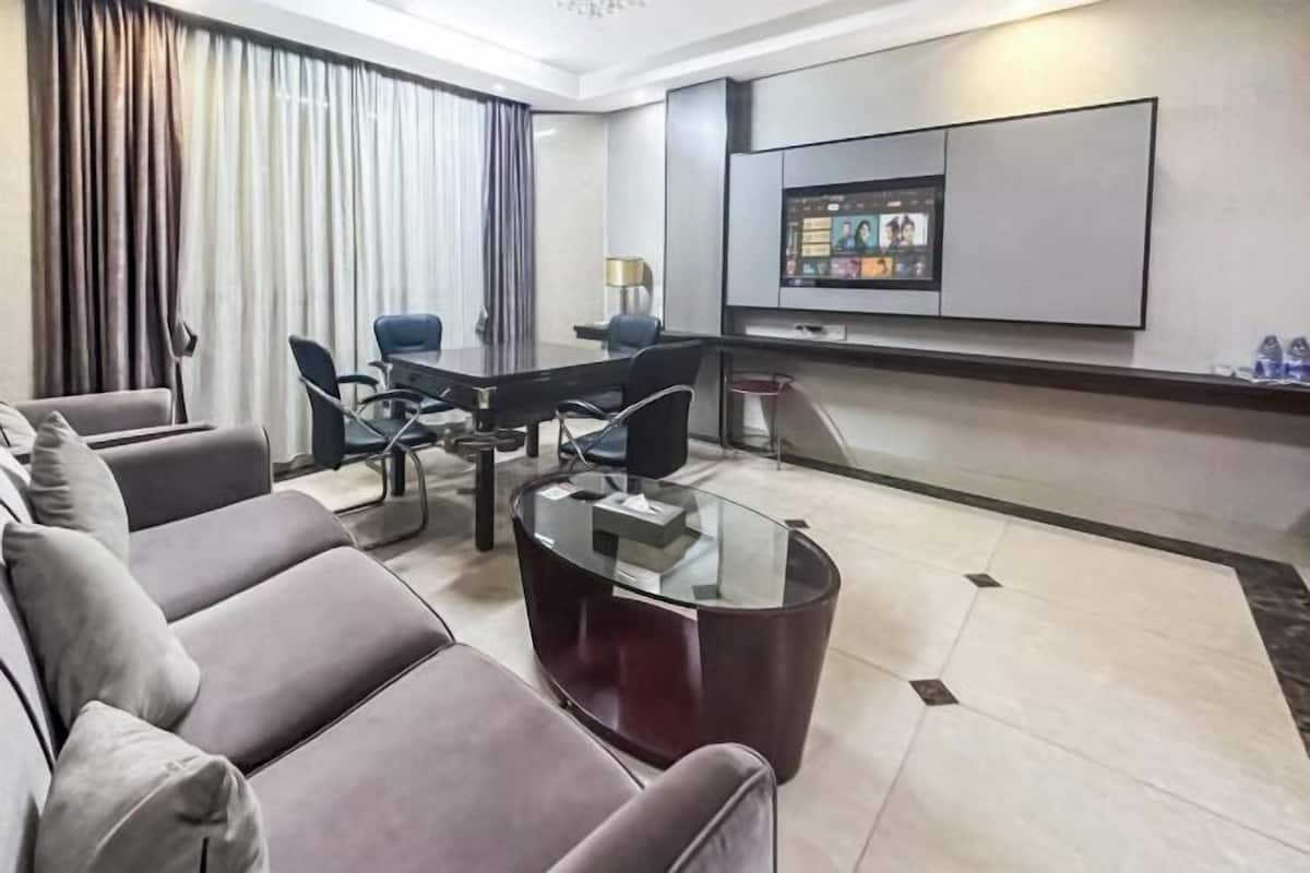 Ein modernes Hotelzimmer mit einer Couch, einem Glastisch, einem Konferenztisch mit Stühlen, einem flachbildfernseher an der Wand und einem großen Fenster mit Vorhängen.