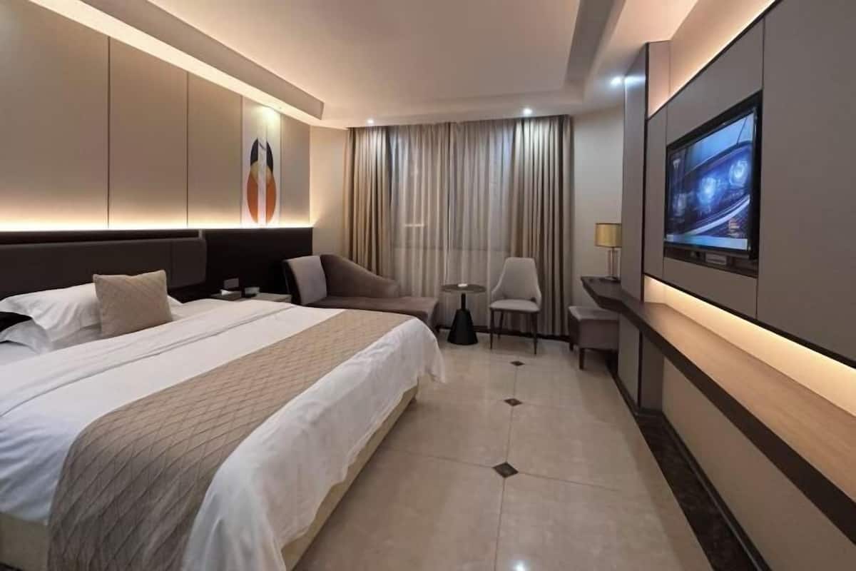 Ein Hotelzimmer mit einem großen Bett, einem an der Wand montierten Fernseher, einer Sitzecke mit einem Sessel und einem kleinen Tisch sowie einem fensterhohen Vorhang.