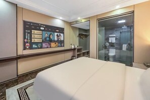 A hotel room with a bed, a wall-mounted TV displaying a video call interface, a glass-enclosed shower, and a small shelf with toiletries. - Yunding Hotel (Huaiji)