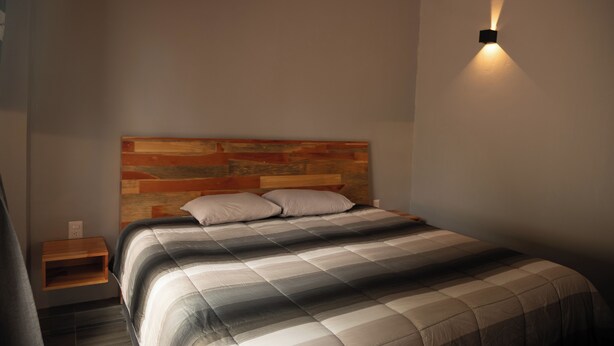 Un dormitorio con cabecera de madera, cama con colcha a rayas, mesita de noche y lámpara de pared.
