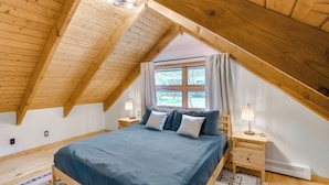 Una camera da letto con soffitto spiovente, un letto con lenzuola blu, due lampade ai comodini, una finestra con tende e un piccolo tavolino di notte.