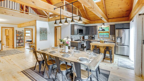 Un'ampia sala da pranzo con soffitto alto e travi in legno, una cucina con elettrodomestici moderni e un angolo salotto accogliente con librerie.