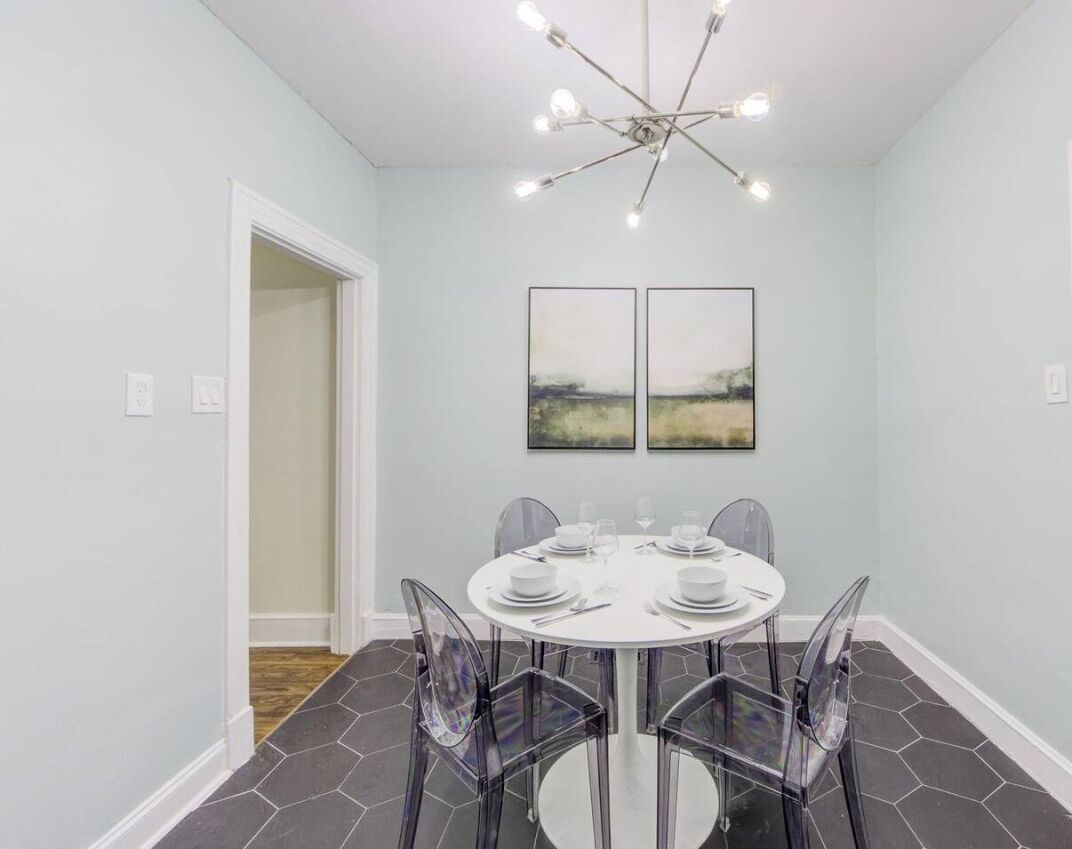 A dining area with a round table, clear chairs, a modern chandelier, and two framed artworks on the wall.