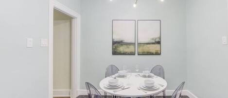 A dining area with a round table, clear chairs, a modern chandelier, and two framed artworks on the wall.