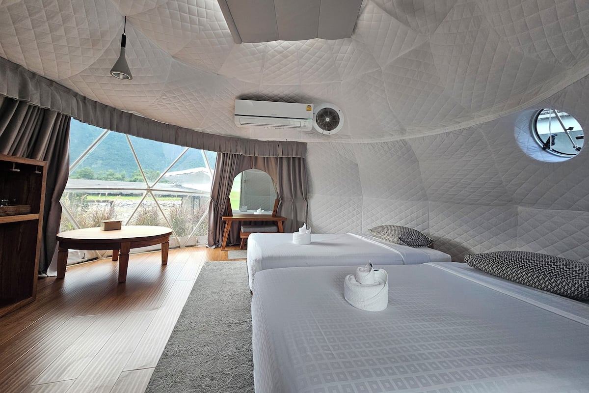 A cozy, modern bedroom with a large bed, a small table, and a chair. It features a large window offering a scenic view.