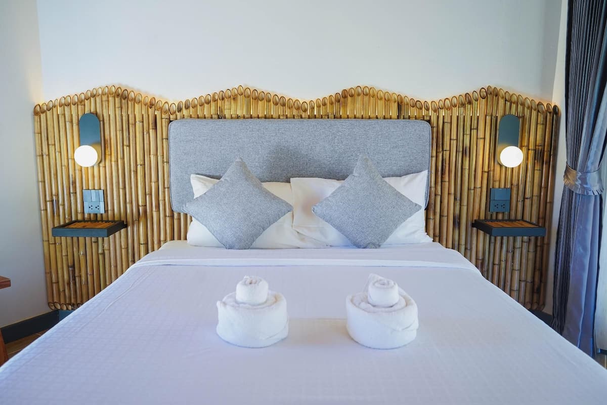 A neatly made bed with two pillows, a bamboo headboard, bedside tables with lamps, and two rolled towels on the bed.
