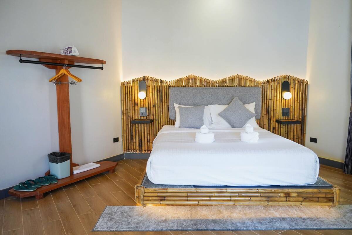 A neatly arranged bedroom with a large bed, bedside lamps, a wooden hanger rack, and a small table with a trash bin.