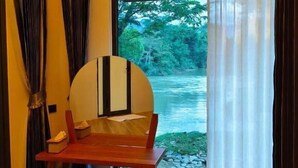 Una habitación con una mesa de madera y un banco, un ventanal con vistas a un río y vegetación, y una cortina parcialmente abierta a un lado.