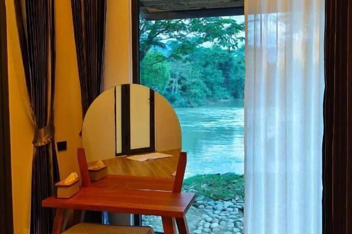 A room with a wooden table and bench, a large window with a view of a river and greenery, and a curtain partially drawn to the side.