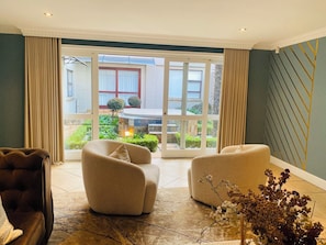 Un salon moderne doté d’une grande porte-fenêtre coulissante donnant sur un jardin, de deux fauteuils beiges, d’un canapé marron foncé et d’un vase contenant des fleurs séchées.