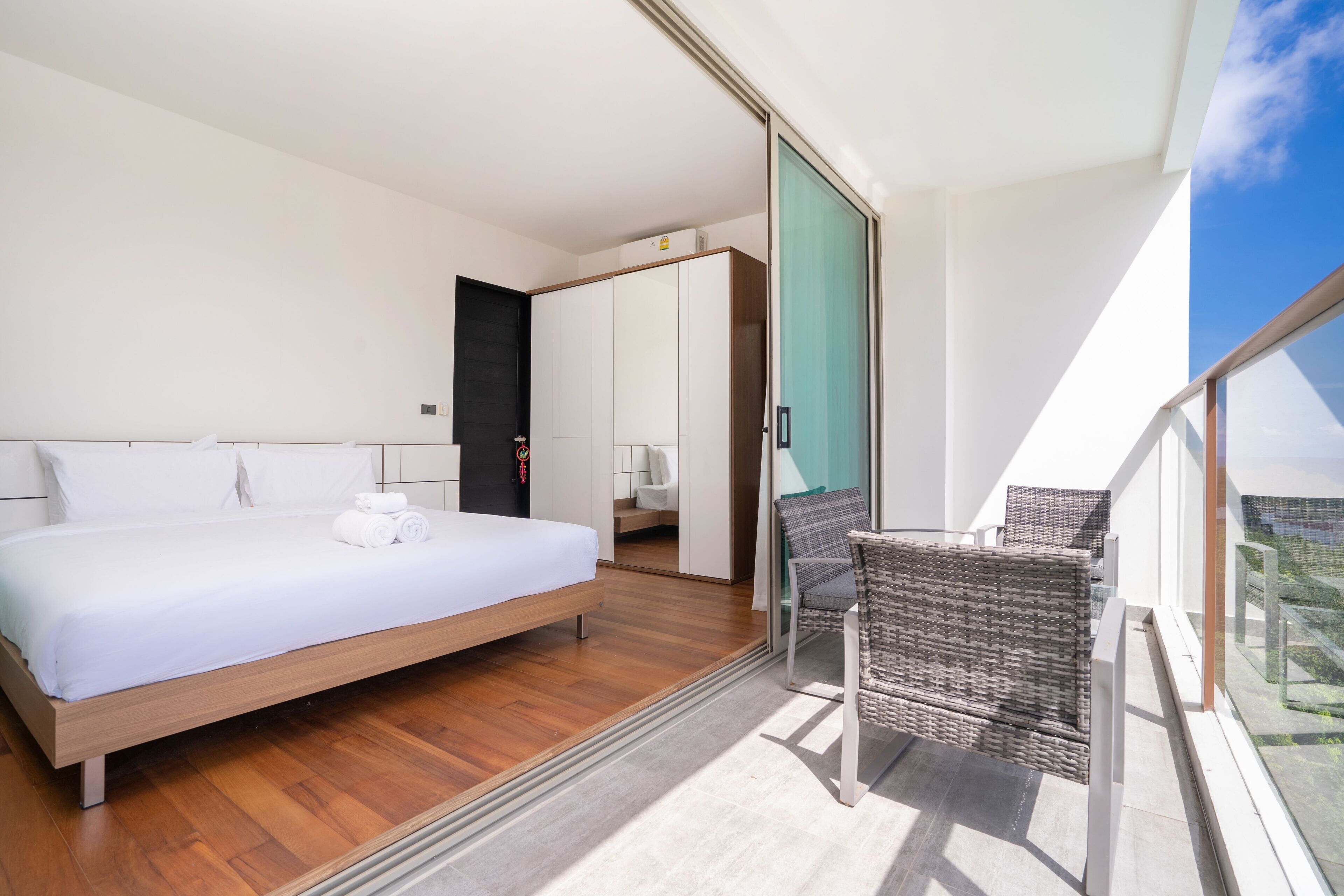 A modern bedroom with a large bed, a balcony with wicker furniture, and a view of the sky.