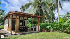 A small wooden structure with a covered patio, surrounded by lush greenery and palm trees.