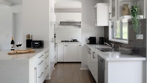A modern kitchen with white cabinetry, a gas stove, a coffee maker, and a window with a plant. - The Village Green Estate Strath Creek (Strath Creek)