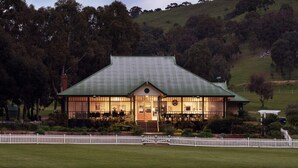 A two-story building with a green roof, surrounded by a white picket fence, set in a hilly landscape with dense trees. - The Village Green Estate Strath Creek (Strath Creek)