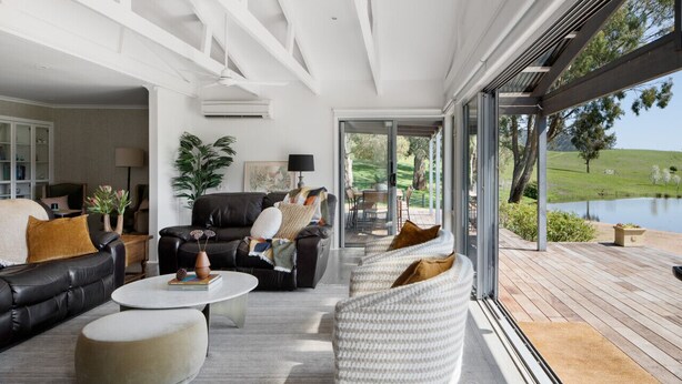 A modern living room with a black leather sofa, white armchairs, and a coffee table. There is a large window providing a view of a lake and greenery. - The Village Green Estate Strath Creek (Strath Creek)