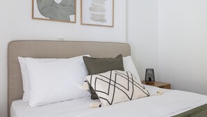 Ein ordentlich bezogenes Bett mit weißen und gemusterten Kissen, einem Kopfteil und einem Nachttischchen mit einer Lampe.