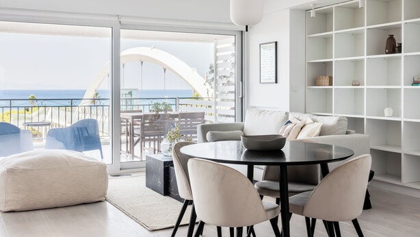 Ein modernes Wohnzimmer mit rundem Tisch, Stühlen, einer Couch und einem großen Fenster mit Meerblick.