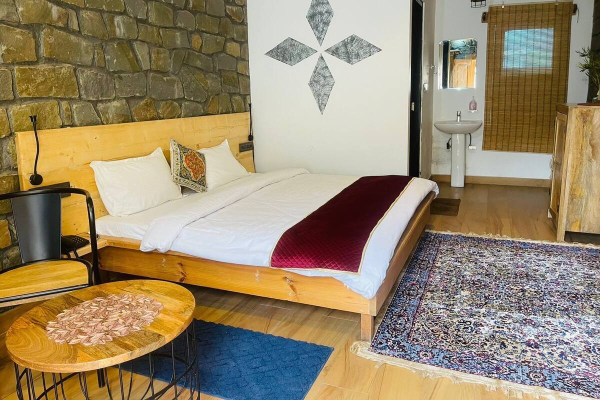Une chambre avec un mur en pierre, un lit avec une literie blanche, une tête de lit en bois, une table ronde en bois agrémentée d’un objet décoratif, un tapis bleu et un tapis à motifs.