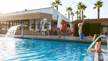 Una piscina amb una cascada, un parasol blanc i palmeres al fons.
