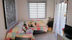A living room with a sofa covered in a colorful quilt, a flat-screen TV on a stand, a decorative wall piece, and a window with blinds. - APT. PLAYAELPORIS (Porís de Abona)