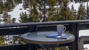 Un tavolo rotondo in vetro con una tazza bianca e un libro, su un balcone affacciato su una montagna innevata e una fitta foresta.