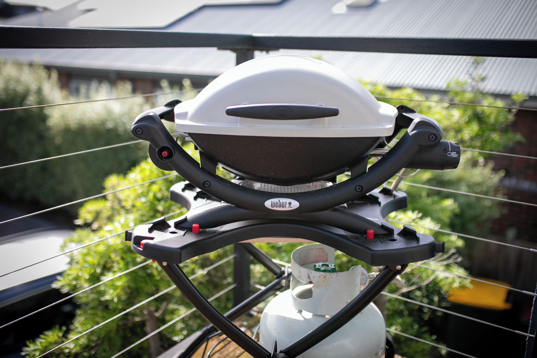 A grill with a white lid and a Weber logo, mounted on a railing with a gas cylinder below.