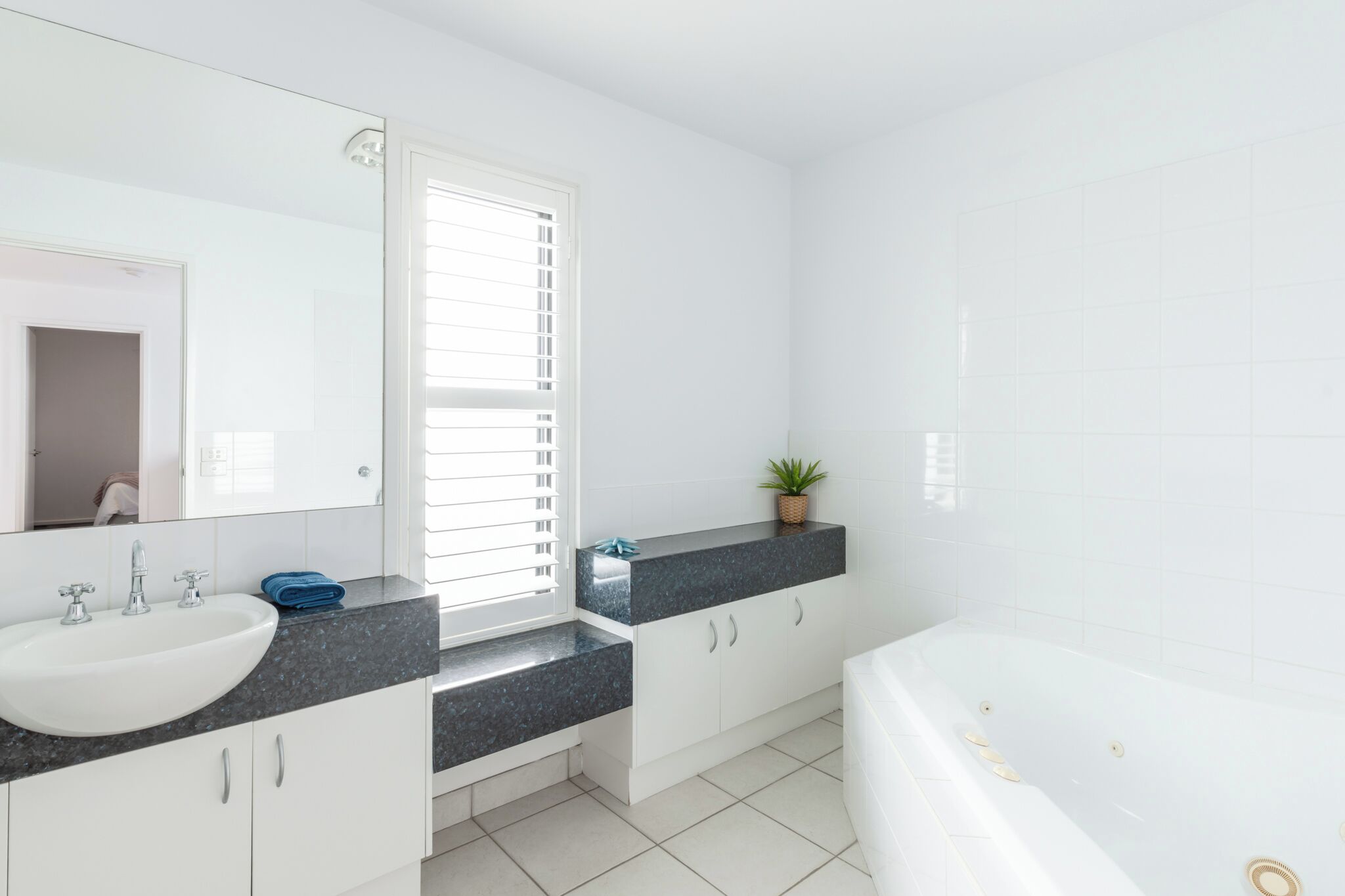 Une salle de bain moderne avec un lavabo blanc, un grand miroir, une baignoire et une fenêtre avec des stores.