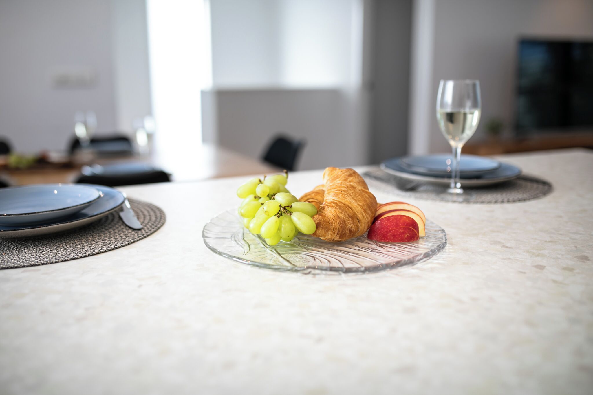 Una mesa de comedor con platos, una copa de vino blanco, uvas y un croissant.