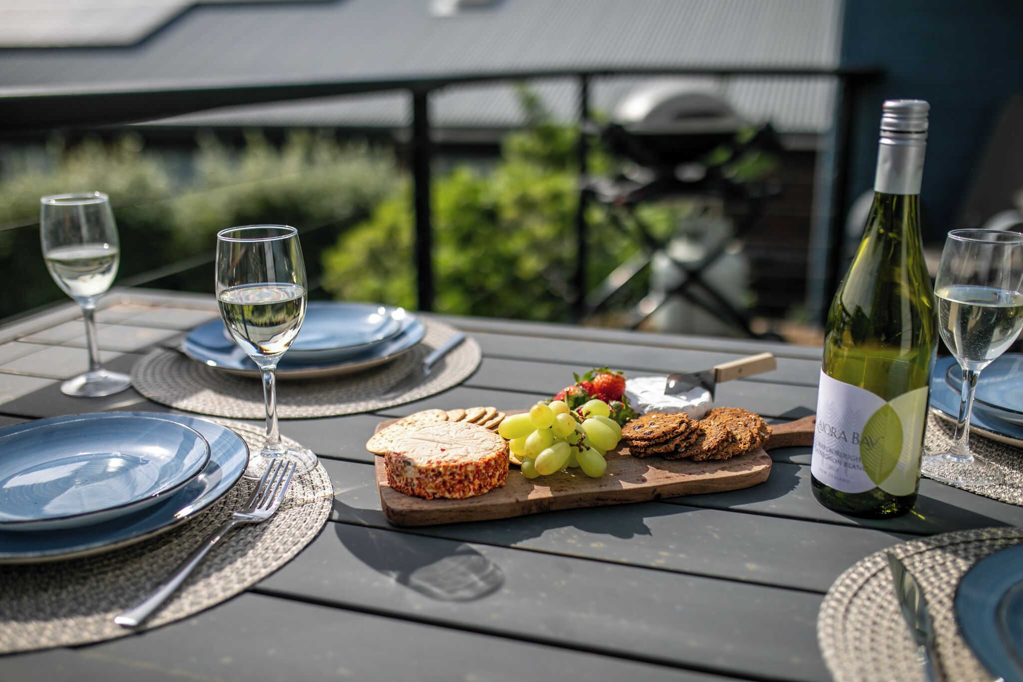 Une table mise pour un repas, avec des assiettes, des couverts, une bouteille de vin et une planche à fromages accompagnée de raisins et de crackers.