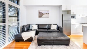 Una sala de estar moderna con un sofá seccional de cuero negro, un manto de pelo blanco y una zona de cocina con armarios blancos y electrodomésticos de acero inoxidable.