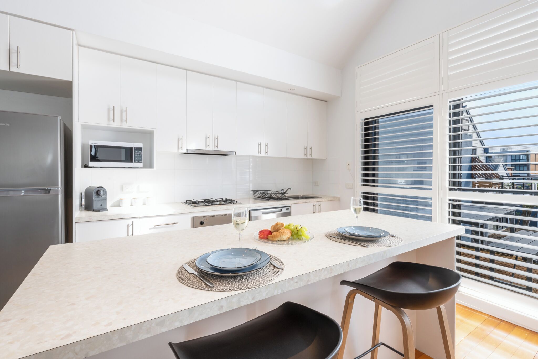 A modern kitchen with white cabinets, a stainless steel refrigerator, a microwave, and a gas stove. There is a dining area with a large island, two black bar stools, and a window with blinds.