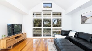 Una sala de estar moderna con piso de madera, un sofá de cuero negro, un mueble de televisión de madera y un ventanal con persianas.