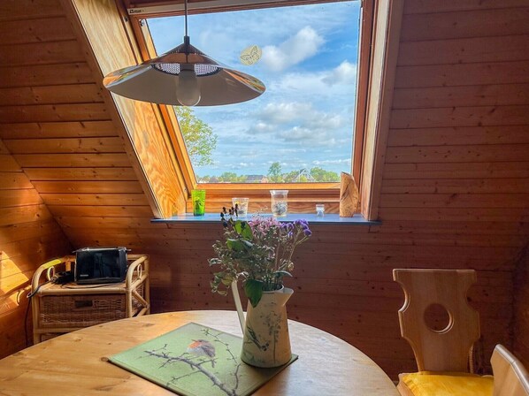 Interior de madeira com uma mesa, um vaso de flores, uma cadeira e uma janela com vista para uma casa.