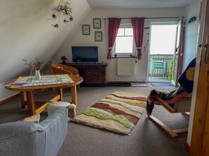 Uma sala de estar acolhedora com uma mesa de madeira, um sofá, uma cadeira de balanço, uma TV e vista para o exterior.
