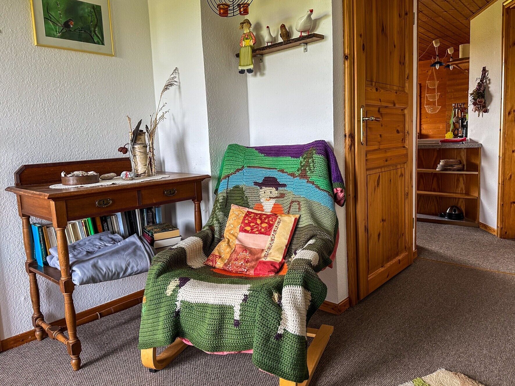 A cozy room with a wooden chair, a desk with books, a shelf with decorative items, and a door leading to another room.